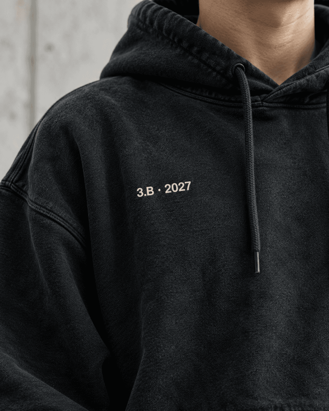 Nærbillede af brystet på en washed black hoodie med en lille printet klassedetalje: 3.B · 2027